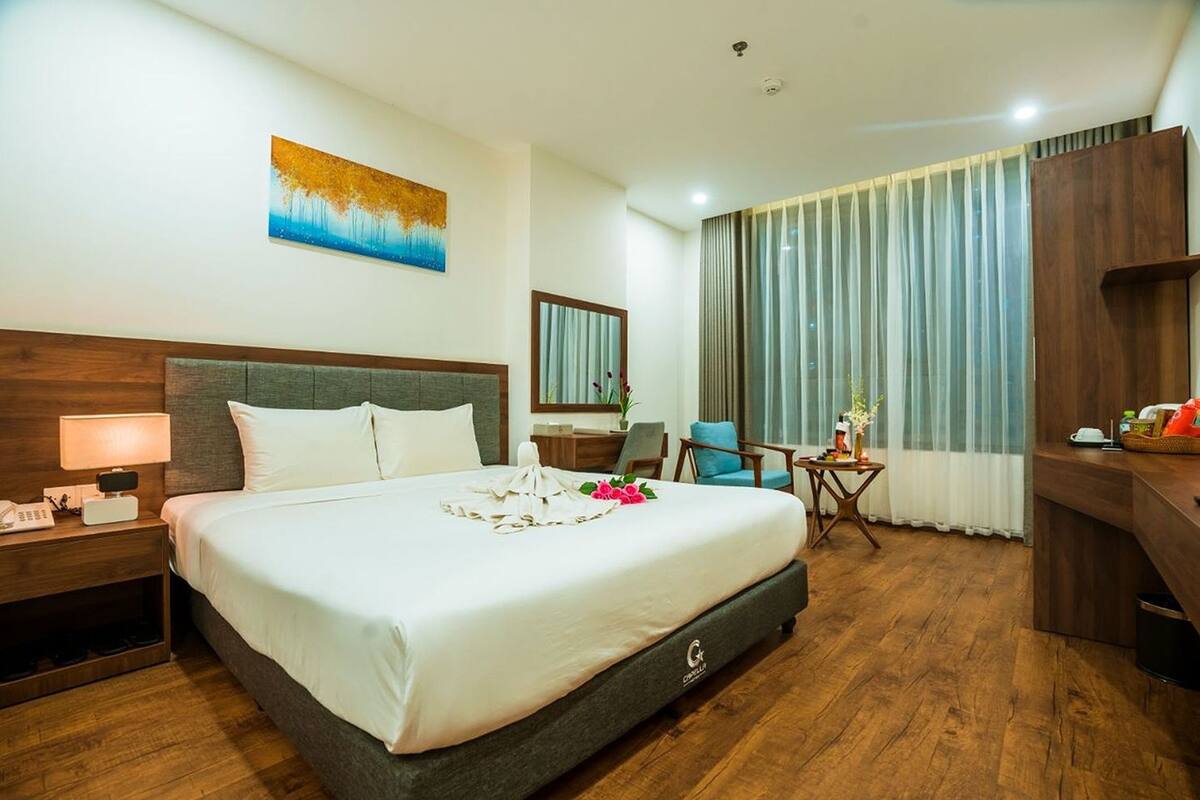 Habitación de hotel con una cama grande, un escritorio, una silla, una mesita, una lámpara, un cuadro en la pared y una ventana con cortinas.