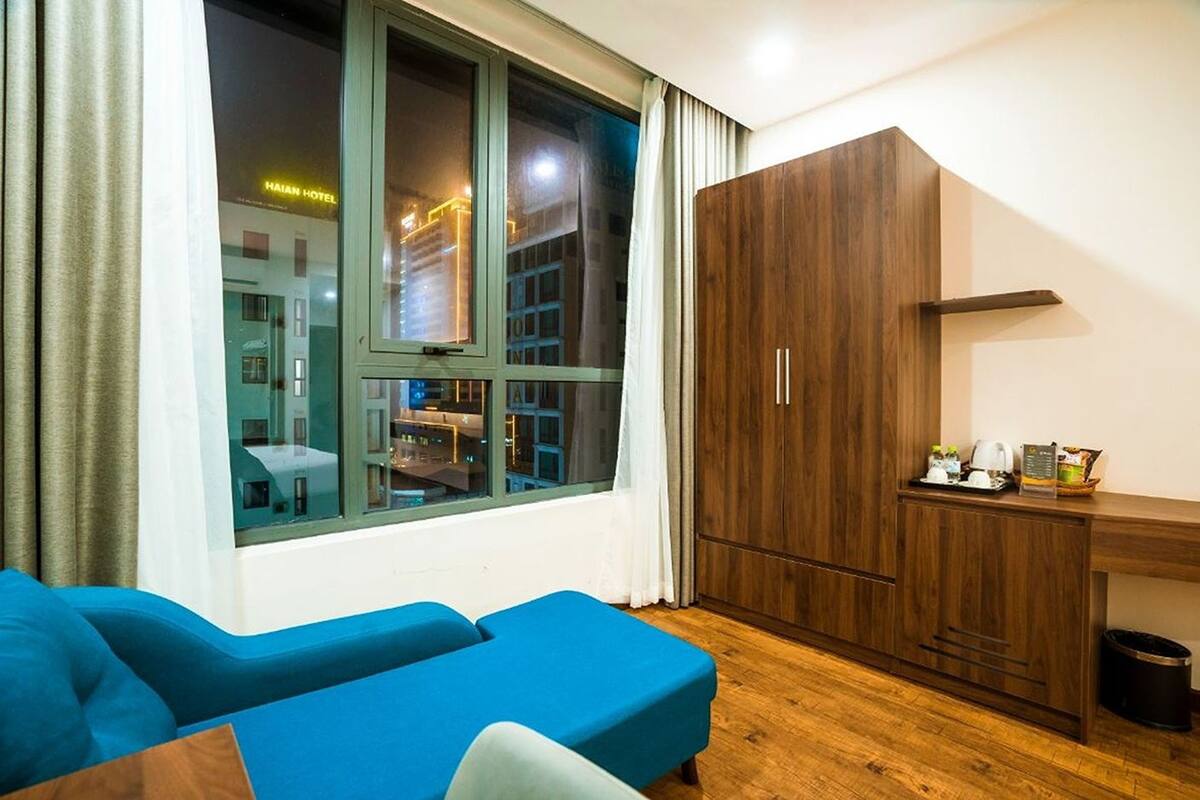 Una habitación moderna con un ventanal grande, un sofá azul, un armario de madera y una mesita con hervidor y tazas.