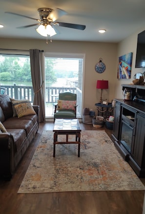 A living room with a ceiling fan, a coffee table, a sofa, a chair, a rug, a TV, a clock, and a view of a lake.