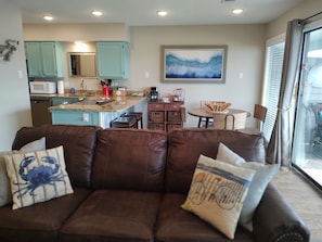 A living room with a brown leather sofa, a kitchen with teal cabinets, and a dining area with a wooden table and chairs.