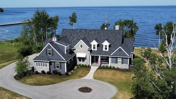 A large, two-story house with a stone facade and dark roofing, situated by a body of water.