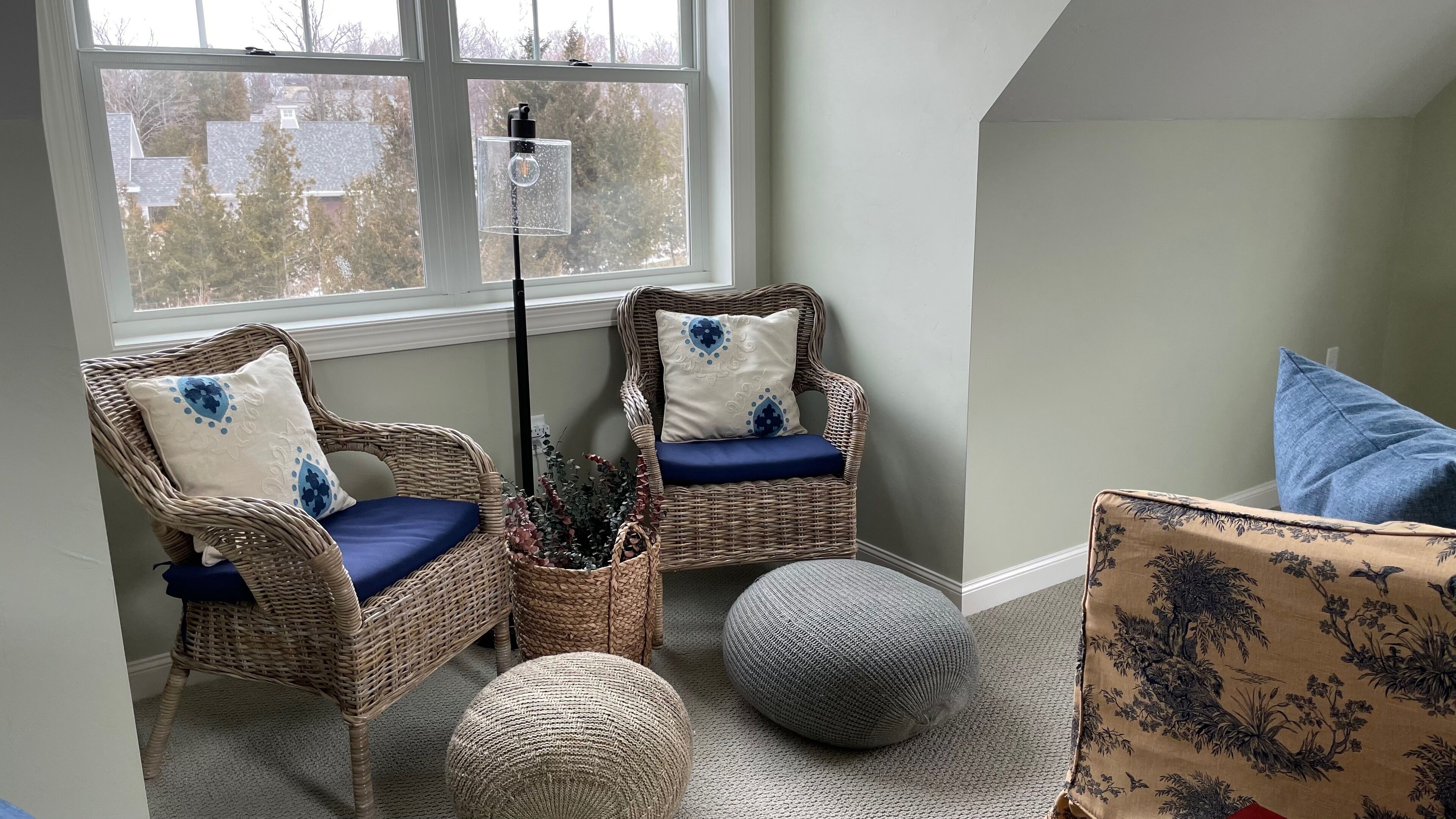 Un coin accueillant avec deux fauteuils en osier, dont l’un est agrémenté d’un coussin bleu, et un petit pouf rond.