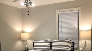 Un lit bien fait, agrémenté d’oreillers à rayures, un ventilateur de plafond et deux lampes de chevet.