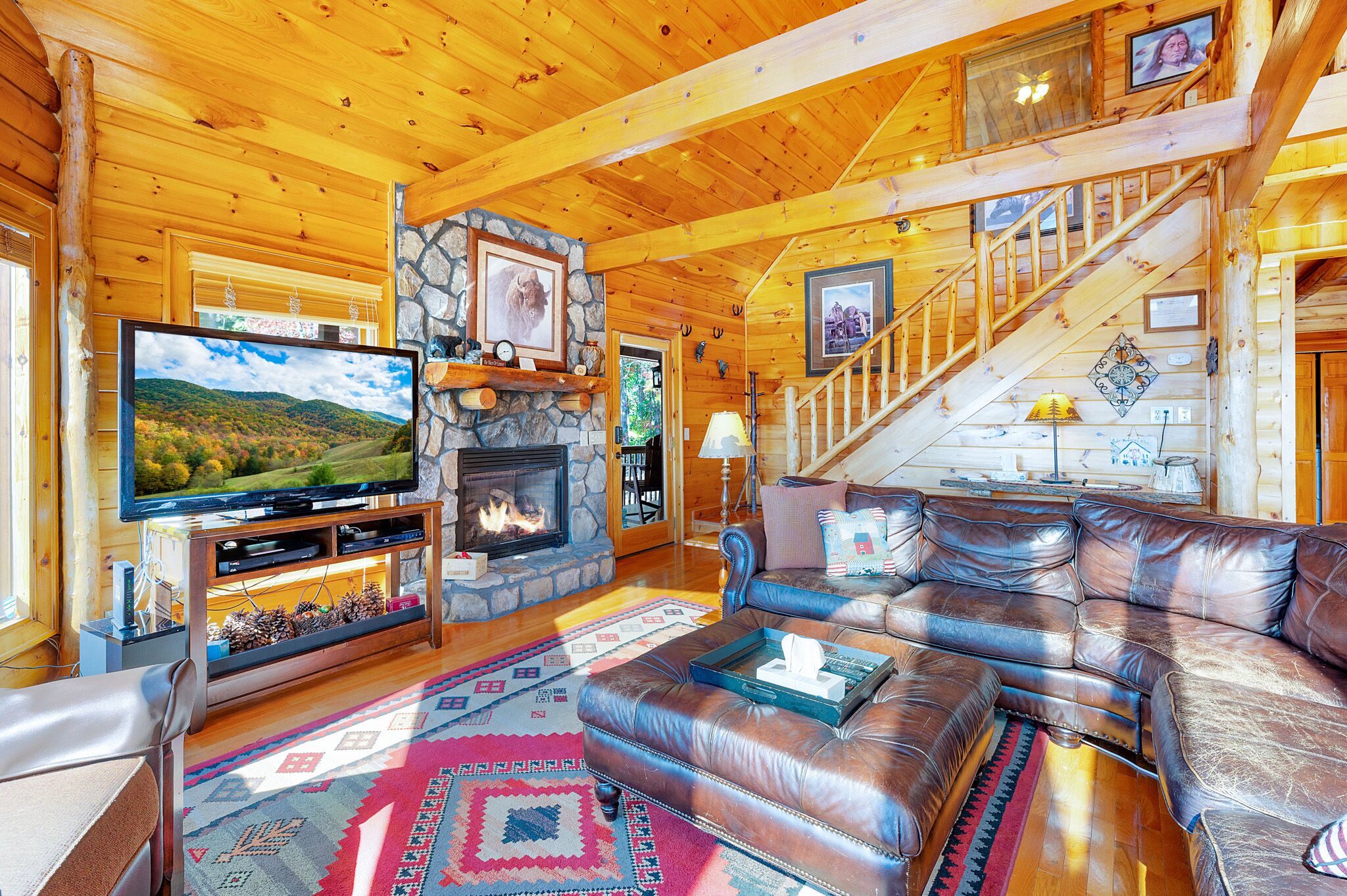 A cozy log cabin interior with a stone fireplace, leather sofa, and a television.