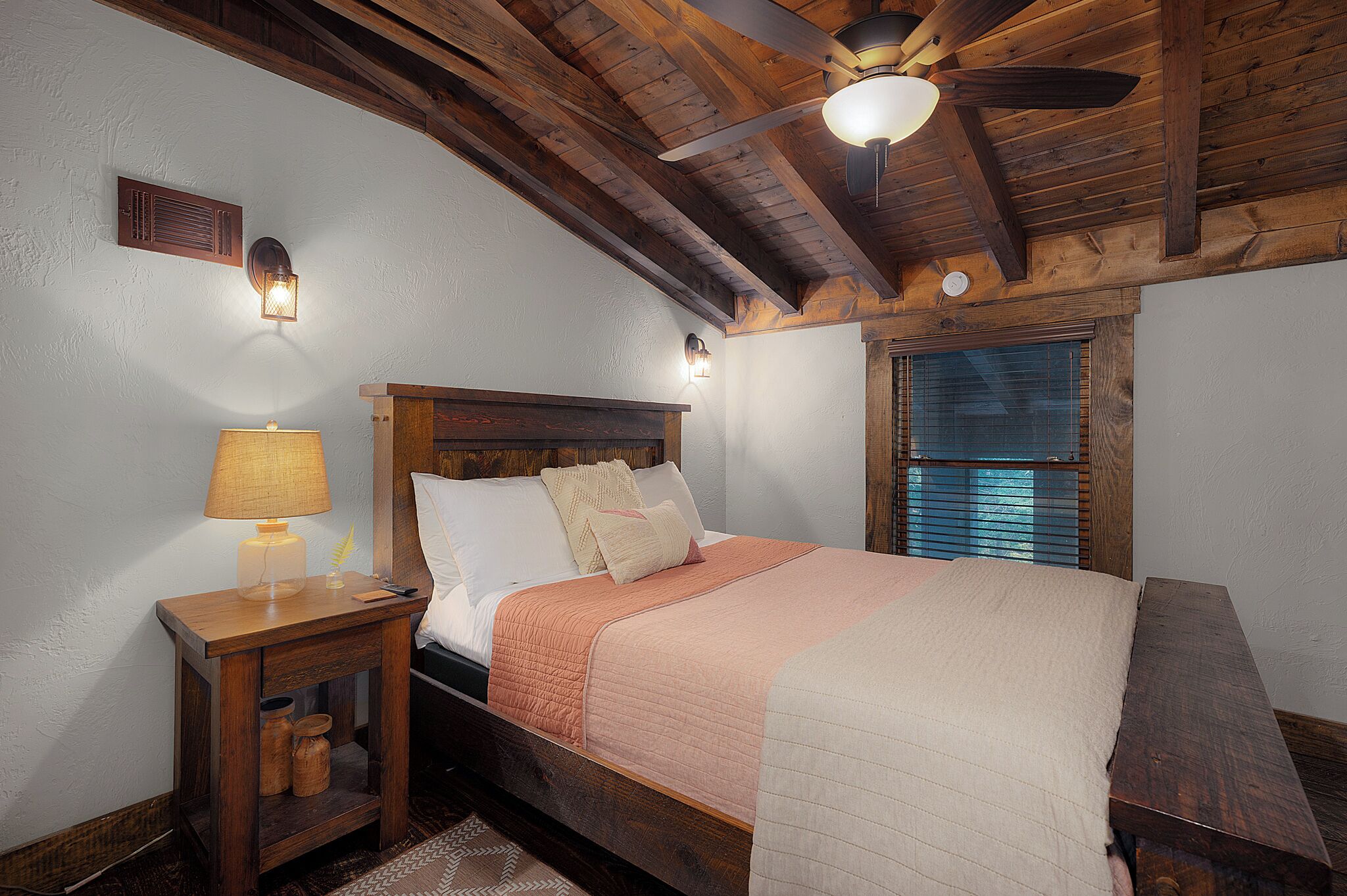 A bedroom with a wooden ceiling, a bed with a headboard, a nightstand with a lamp, and a window with blinds.