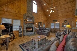 A spacious log cabin interior with a stone fireplace, leather sofas, and a dining area.
