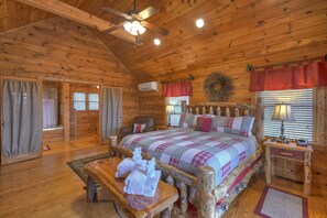 A log cabin bedroom with a large bed, wooden furniture, a ceiling fan, and a window with blinds.
