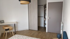 Quarto com piso de madeira, escrivaninha branca, cadeira, cama com colcha estampada, guarda-roupa e porta.