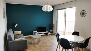 Uma sala de estar moderna com uma parede de destaque azul-esverdeado, um sofá cinza, um rack branco para televisão e uma área de jantar com mesa e cadeiras redondas.