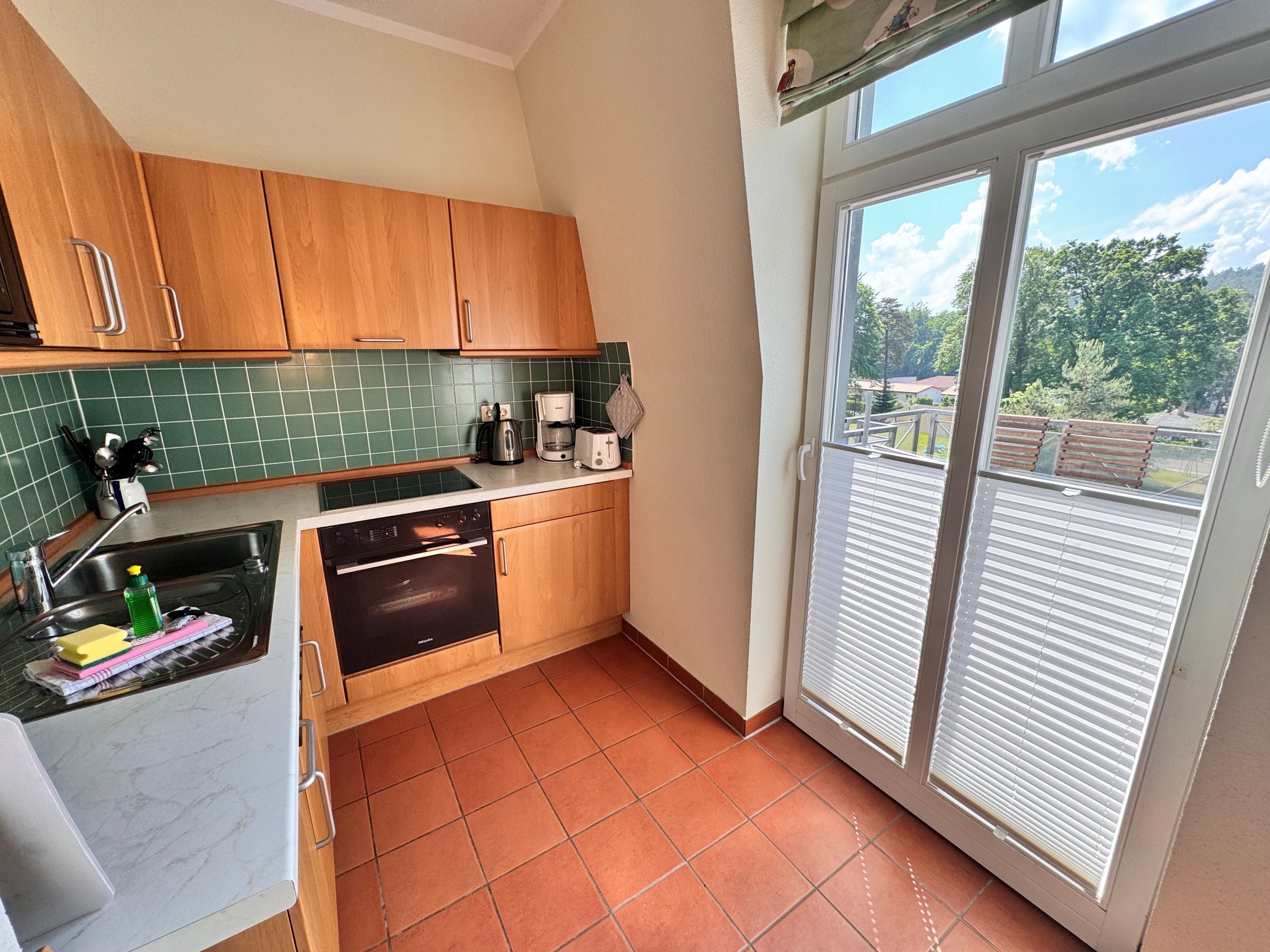 A kitchen with a stove, oven, and coffee maker, a window with blinds, and a view of greenery outside.