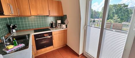 A kitchen with a stove, oven, and coffee maker, a window with blinds, and a view of greenery outside.
