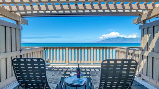 Un balcón con dos sillas y una mesa, con vistas a una playa de aguas azules cristalinas y montañas distantes.