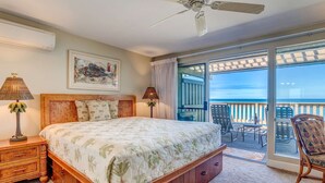 Un dormitorio con una cama grande, un ventilador de techo, una mesita de noche con lámpara, un cuadro en la pared y un balcón con mobiliario exterior.
