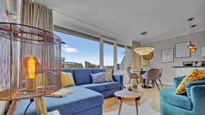 A modern living room with a blue sofa, a round coffee table, and a hanging light fixture. - Seaside SA 140 - Seaside, 2 Zi, 65m², Balkon, Meerblick, 2. OG Fahrstuhl (Scharbeutz)
