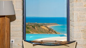 Una habitación con pared de piedra, una silla de madera, una lámpara y vista al océano a través de un ventanal arqueado.