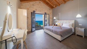 Un dormitorio con cama, mesita de noche y escritorio con espejo. Se ve el mar a través de un ventanal de vidrio.