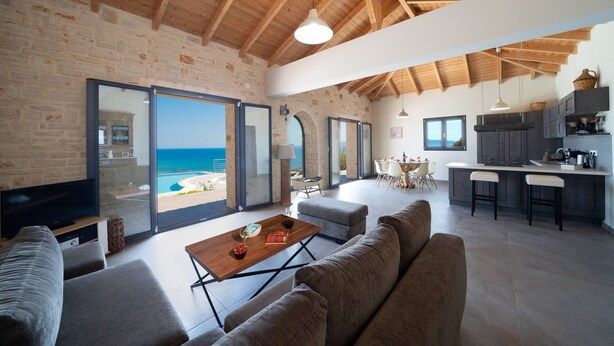 Un salón amplio con televisor grande, asientos cómodos, techo de madera y vista al océano a través de puertas de cristal.