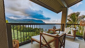 A balcony with a wooden chair and table, a wicker basket, and a view of mountains and greenery. - Hanalei Bay Resort 9305 (Hanalei Bay Hideaway) AC! (Princeville)