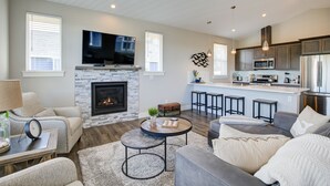 A modern living room with a fireplace, a flat-screen TV, and a kitchen area with bar stools. - Tranquility Base Cottage - Oyhut Bay Seaside Village (Ocean Shores)
