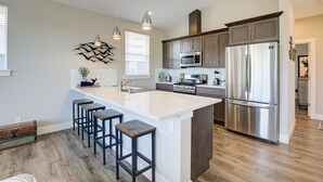 A modern kitchen with a large island, stainless steel appliances, and bar stools. - Tranquility Base Cottage - Oyhut Bay Seaside Village (Ocean Shores)