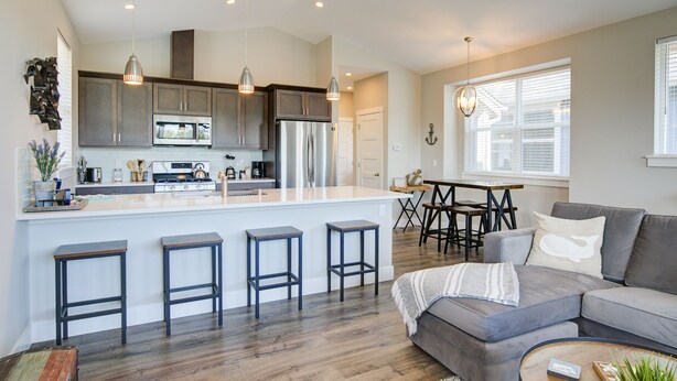 A modern kitchen with a central island, bar stools, a dining area with a table and chairs, and a living room with a sofa and coffee table. - Tranquility Base Cottage - Oyhut Bay Seaside Village (Ocean Shores)
