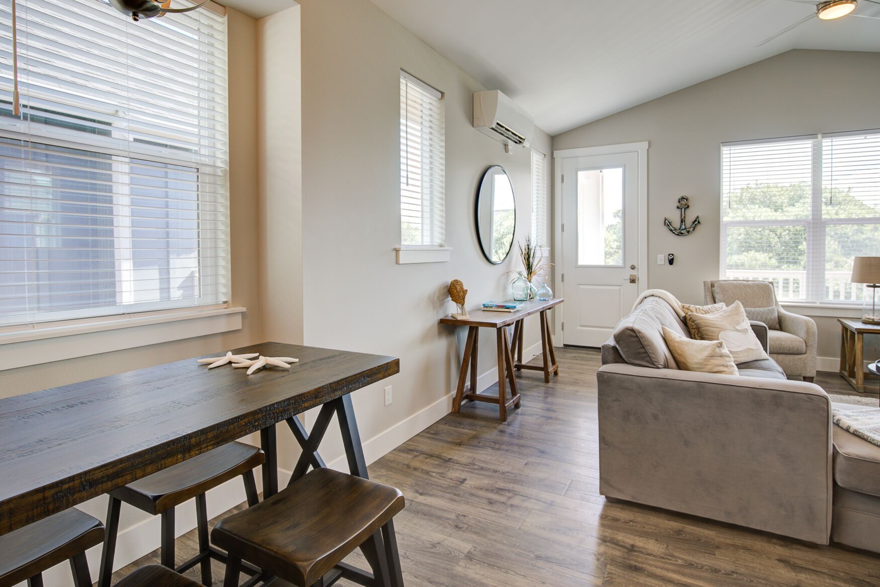 A spacious living room with a wooden dining table, a sofa, and a coffee table. There are windows with blinds, a door, and an air conditioning unit on the wall.