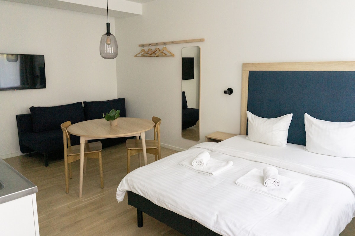 Quarto de hotel com uma cama, uma mesa redonda, duas cadeiras, uma mesinha, um espelho e um candeeiro suspenso.