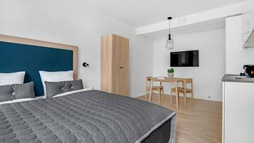 A modern bedroom with a large bed, a wooden wardrobe, a small dining table with chairs, and a TV mounted on the wall.