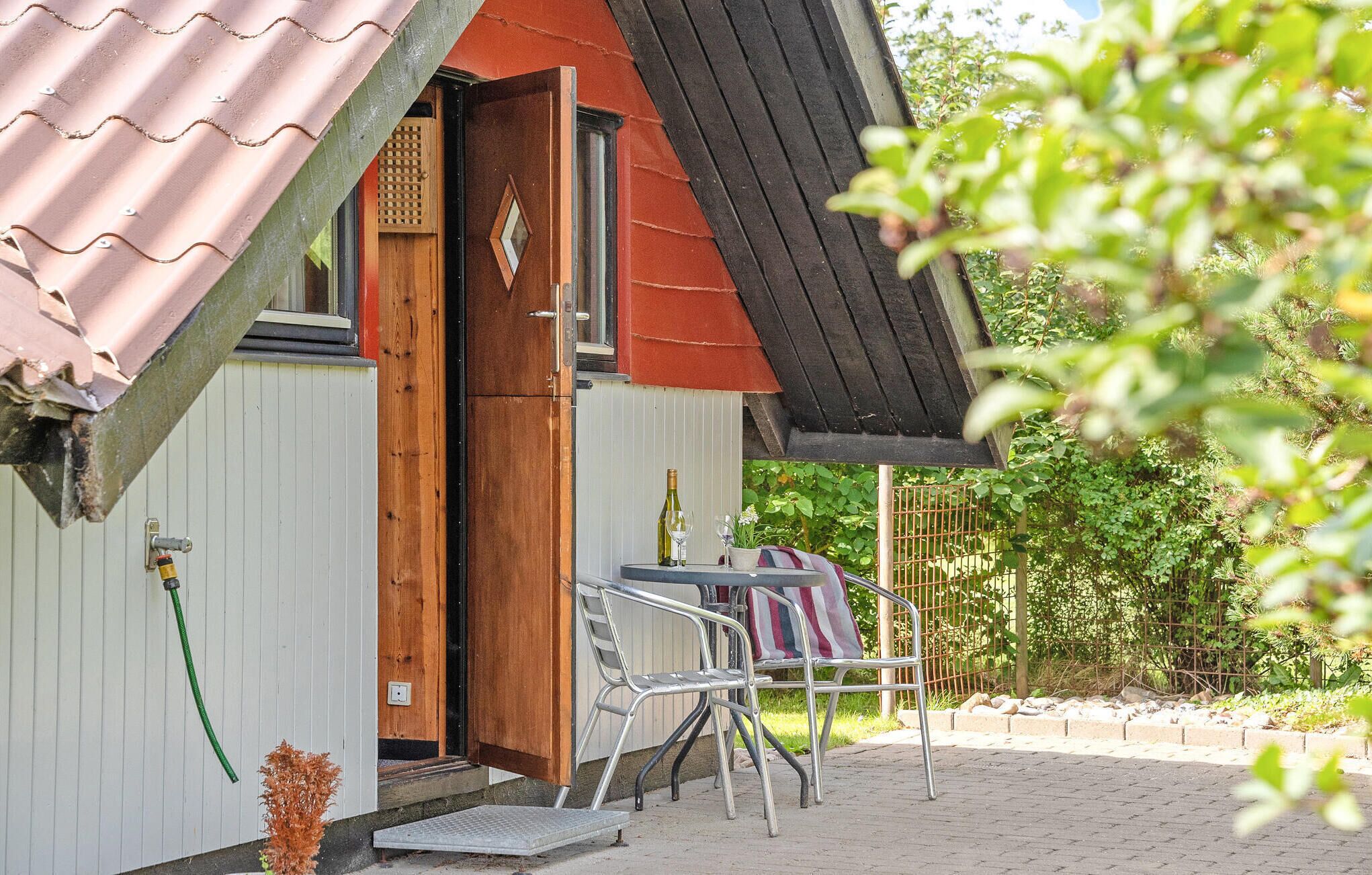 Ein kleines Haus mit rotem Dach und einer Holztür. Vor der Tür steht ein Tisch mit Stühlen, und im Hintergrund ist ein Garten mit Pflanzen und einem Zaun zu sehen.