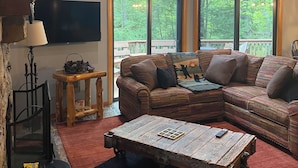A cozy living room with a large sectional sofa, a wooden coffee table, and a fireplace. There are large windows offering a view of the outdoors. - charming chalet in the Lake Naomi community (Pocono Pines)