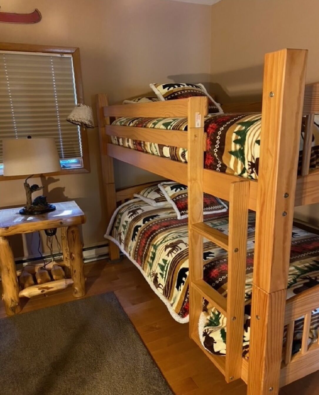 Un lit superposé avec une couvre-lit à motifs, un petit bureau en bois avec une lampe et une fenêtre avec des stores.