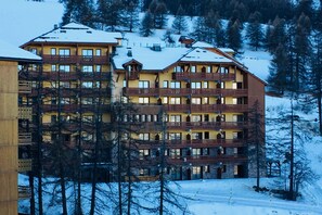 A multi-story hotel with balconies, surrounded by snow-covered trees and a mountainous backdrop. - Risoul - Studio 406 - Pool/Sauna - Fully equipped - WIFI - Foot of slopes (Risoul)