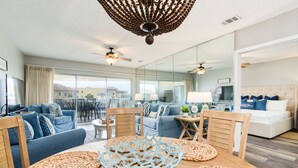 A spacious living room with a large window, a ceiling fan, and a dining area with a wooden table and woven placemats.