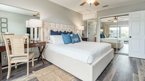 A bedroom with a large bed, a desk, a chair, a ceiling fan, and a view of the outside.