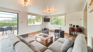 A spacious living room with a large sofa, two armchairs, a wooden coffee table, and a TV mounted on the wall. There are large windows providing views of greenery outside. - Gorgeous home in Ledinghem with sauna (Ledinghem)