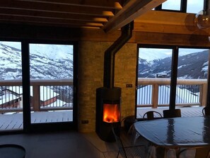 Una habitación acogedora con estufa de leña, ventanas amplias con vistas a una montaña nevada y un comedor.