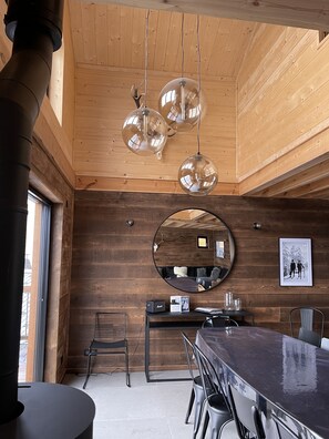 Un interior moderno con techo de madera, una zona de comedor con una mesa grande y sillas, un espejo redondo y un cuadro en la pared.