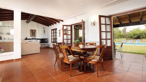 A spacious dining area with a round table and six chairs, a kitchen in the background, and a patio with a pool view. - COLLET VELL. Lovely stone-clad villa with large garden (Pollensa)