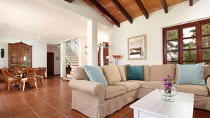 A spacious living room with a beige sectional sofa, a dining area with wooden chairs, a staircase, and a large window with a view of trees. - COLLET VELL. Lovely stone-clad villa with large garden (Pollensa)