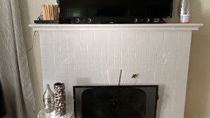 Un salon avec une cheminée blanche, un téléviseur à écran plat fixé au-dessus, et une petite table agrémentée d’objets décoratifs.