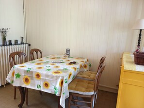 Ein Essbereich mit Tisch, bedeckt mit einer Tischdecke mit Sonnenblumenmuster, zwei Holzstühlen, einem kleinen dekorativen Gegenstand auf dem Tisch und einem gelben Schrank mit Lampe darauf.