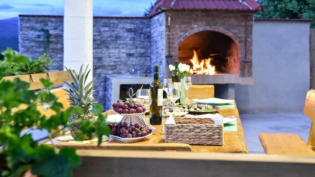 Un'area pranzo all'aperto con un forno a mattoni, un tavolo di legno apparecchiato con cibo e bevande, e un muro in pietra sullo sfondo.