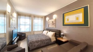 A modern bedroom with a large bed, a desk, a chair, and a sofa. There are windows with blinds, a painting on the wall, and a lamp on the desk. - Suite Central Station Milano (Milano)
