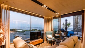 Ein modernes Wohnzimmer mit großem Fernseher, bequemen Sitzmöglichkeiten und Blick auf Stadt und Wasser.