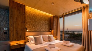 Ein modernes Hotelzimmer mit einem großen Bett, einem Holzkopfende und Blick auf die Stadt durch große Fenster.