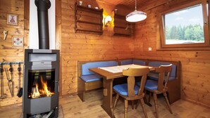 A cozy cabin interior with a wood-burning stove, a dining table with blue upholstered chairs, and a window overlooking a forest.