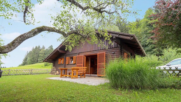 A rustic wooden cabin with an open door, a wooden bench outside, and a car parked nearby.