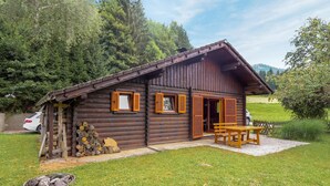 A rustic wooden cabin with a patio, a stack of firewood, and a car parked nearby.