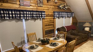Un coin repas chaleureux avec une table et des chaises en bois, entouré de murs en bois et agrémenté d’une fenêtre avec des stores.