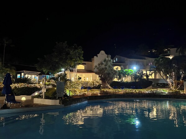Area piscina di notte con un edificio sullo sfondo, illuminata da luci a stringa.